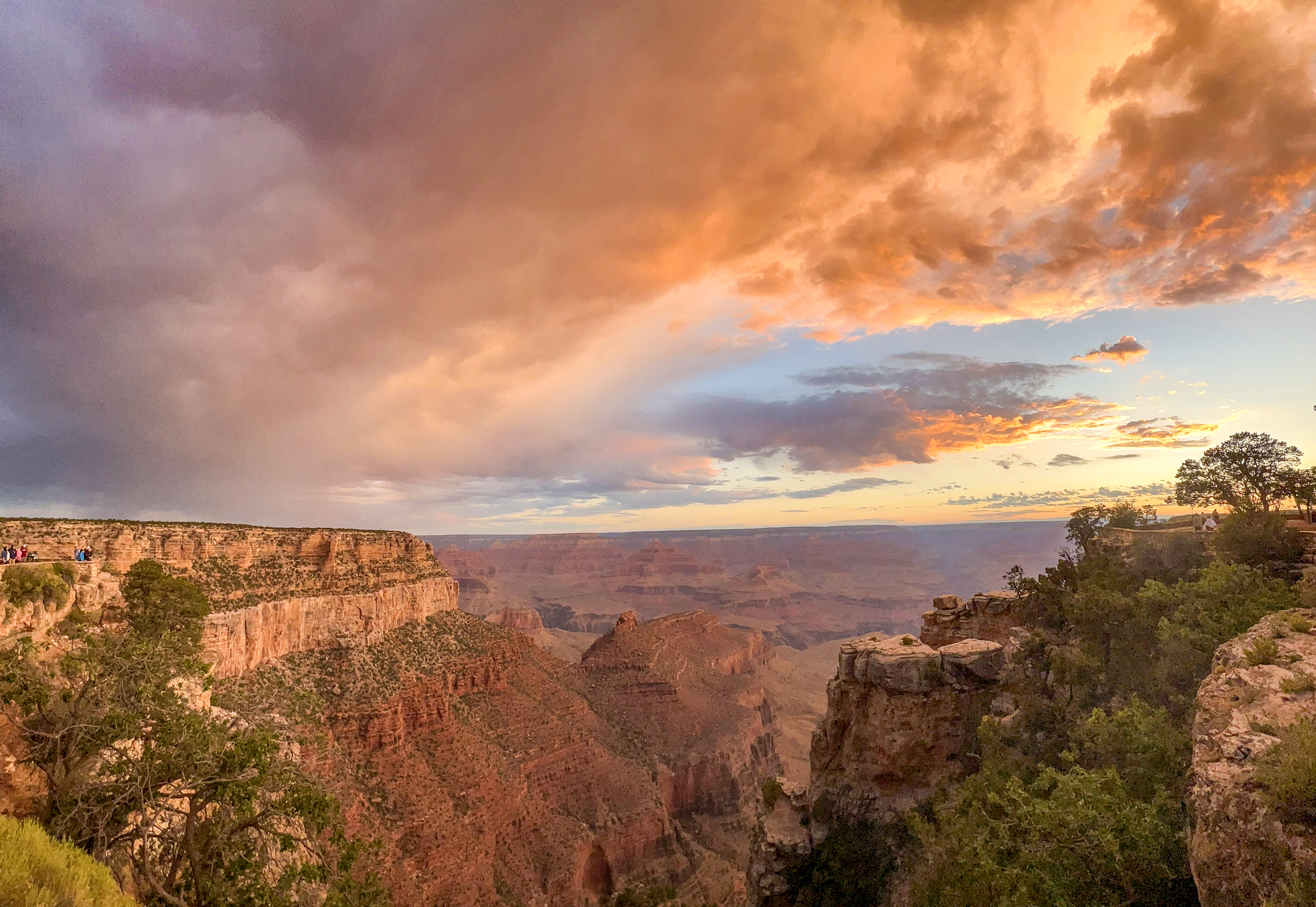 As the sun is rising, it illuminates summer monsoon clouds with brilliant orange light which reflects downward onto the cliffs, peaks and rock formations within a vast mile deep canyon.