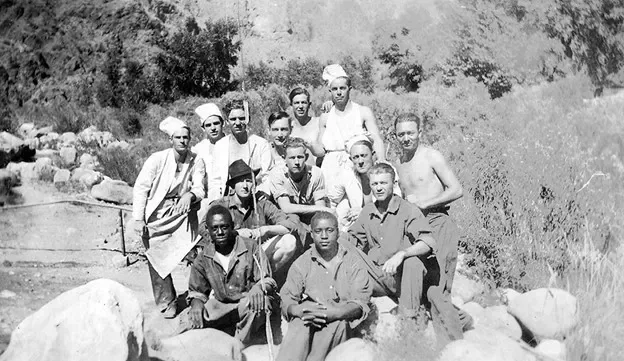 Men of different races sit on rocks in a stream, some with chefs hats on.