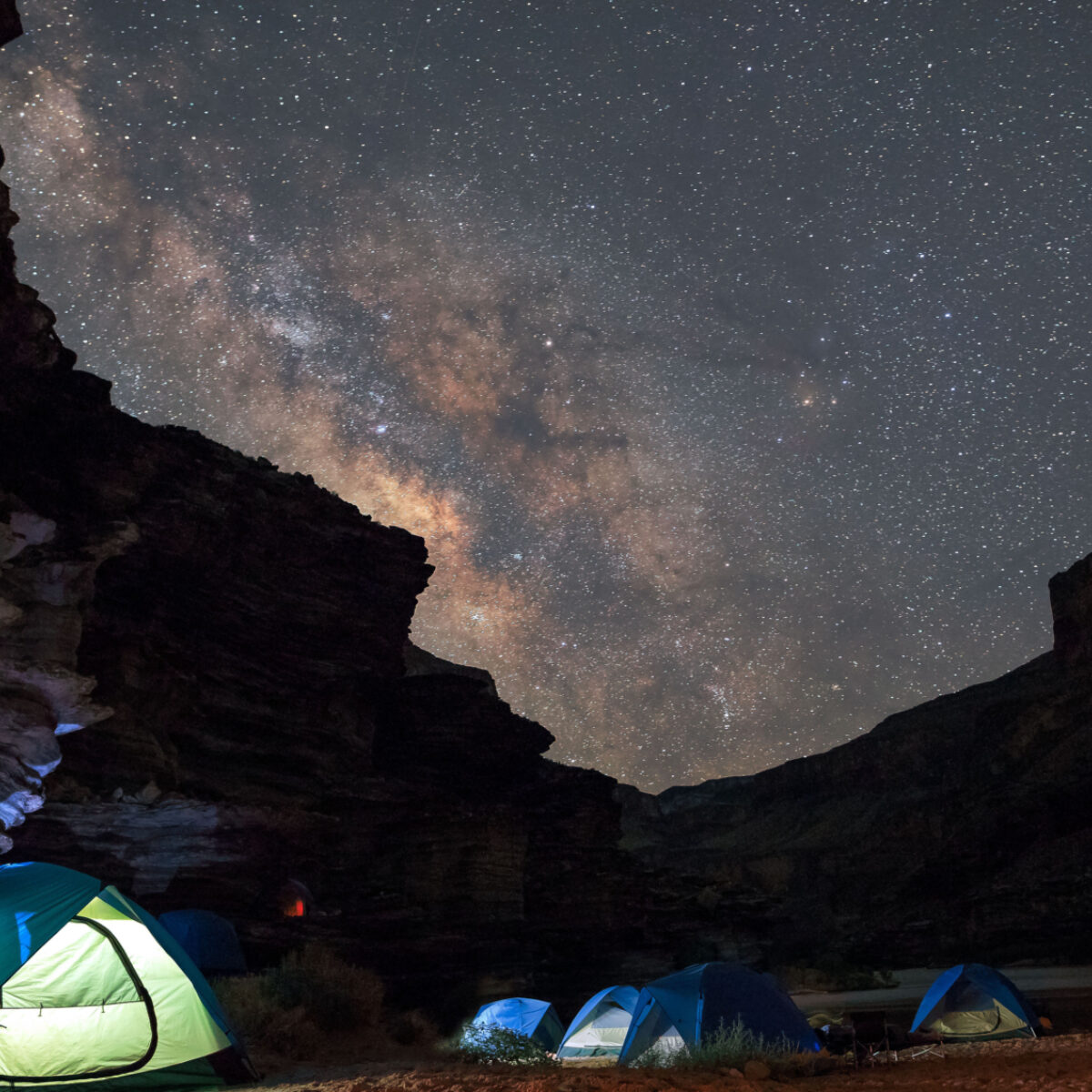 Photo with tents in foreground lit up and the milky way up high. Campers are at the bottom of Grand Canyon camping.