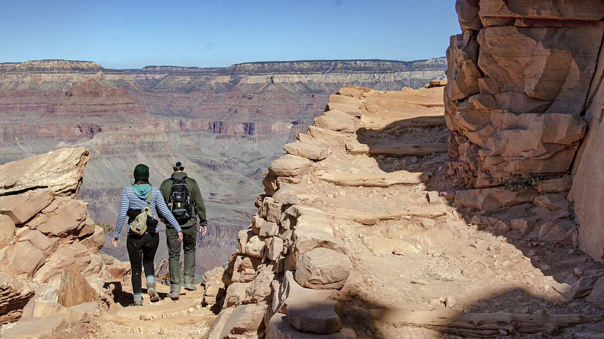 2 day hikers descending have just turned the corner of a rugged backcountry trail switchback built into a cliff. NPS/M.Quinn