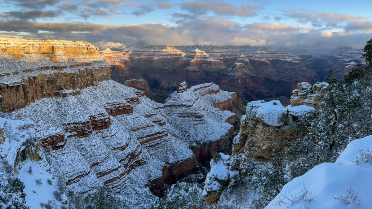 Snow cover extending down several thousand feet of cliffs and slopes into a vast, mile deep canyon landscape, with patchy clouds across the sunrise sky and snowy peaks in the distance.