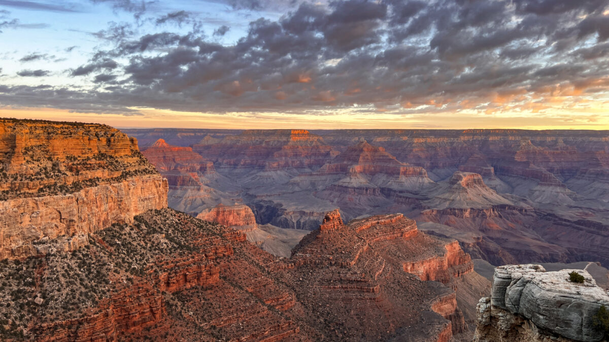Orange sunrise light beginning to illuminate the peaks, cliffs and rock formations within a vast canyon landscape. A patchwork of dark clouds stretches across the sky, their undersides partially tinged by the orange light.