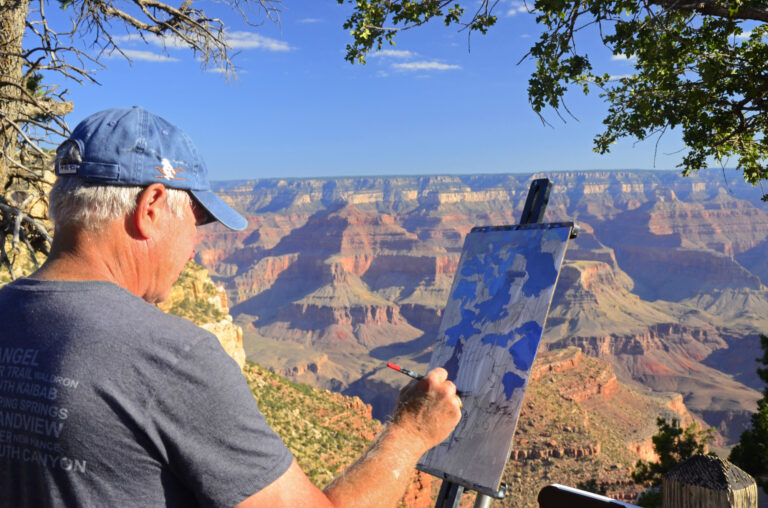 Artist in blue baseball hat drawing canyon view while standing on canyon edge during evening sun at Grand Canyon National Park Celebration of Art Event