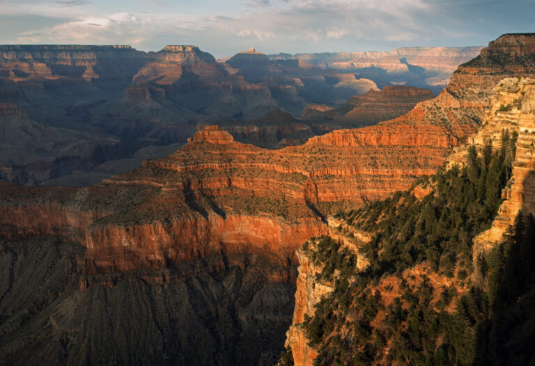 Looking down into a vast canyon landscape mostly in shadow the foreground several ridgelines are illuminated by a golden orange light NPS Michael Quinn