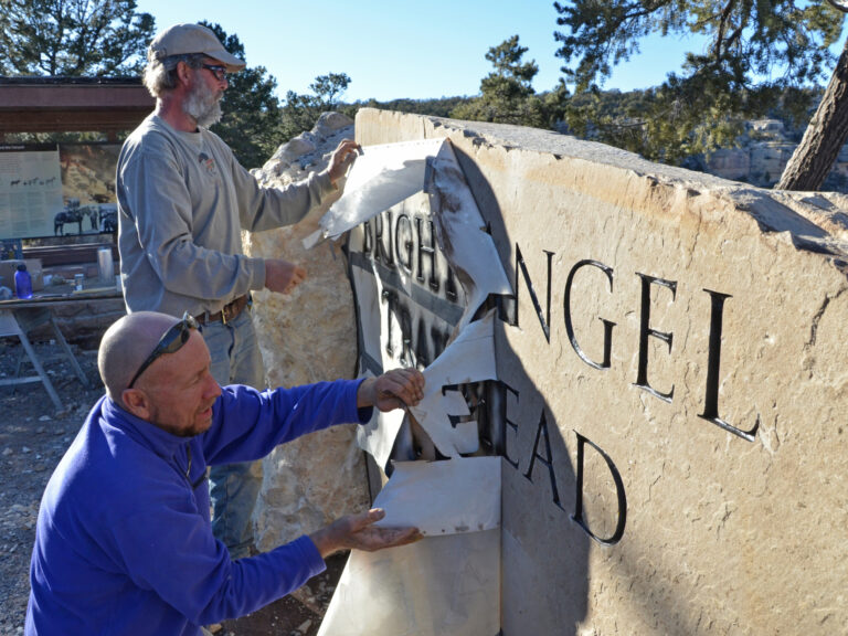 Grand Canyon National Park: Bright Angel Trailhead Sign 2863
March 1, 2013, After sandblasting and staining the letters, stone cutter Andy Dufford (front) and stone mason Brad Quinn remove the stencil from the new Bright Angel Trailhead Sign. The sign consists of a 9’x5’x12” sandstone slab weighing approximately 3 tons with two ‘bookend’ boulders on either side. Each of the bookend boulders was sawn and chiseled to create a slot to help support the slab vertically. The boulder on the east side also serves as a bench. The sign is also supported by a concrete foundation. “Bright Angel Trailhead” is sandblasted into the slab. It’s sited to capture a background view of the canyon, and has space behind for hikers to pose for a photo op. The ID sign was created by Chevo Studios. NPS photo by Michael Quinn.
