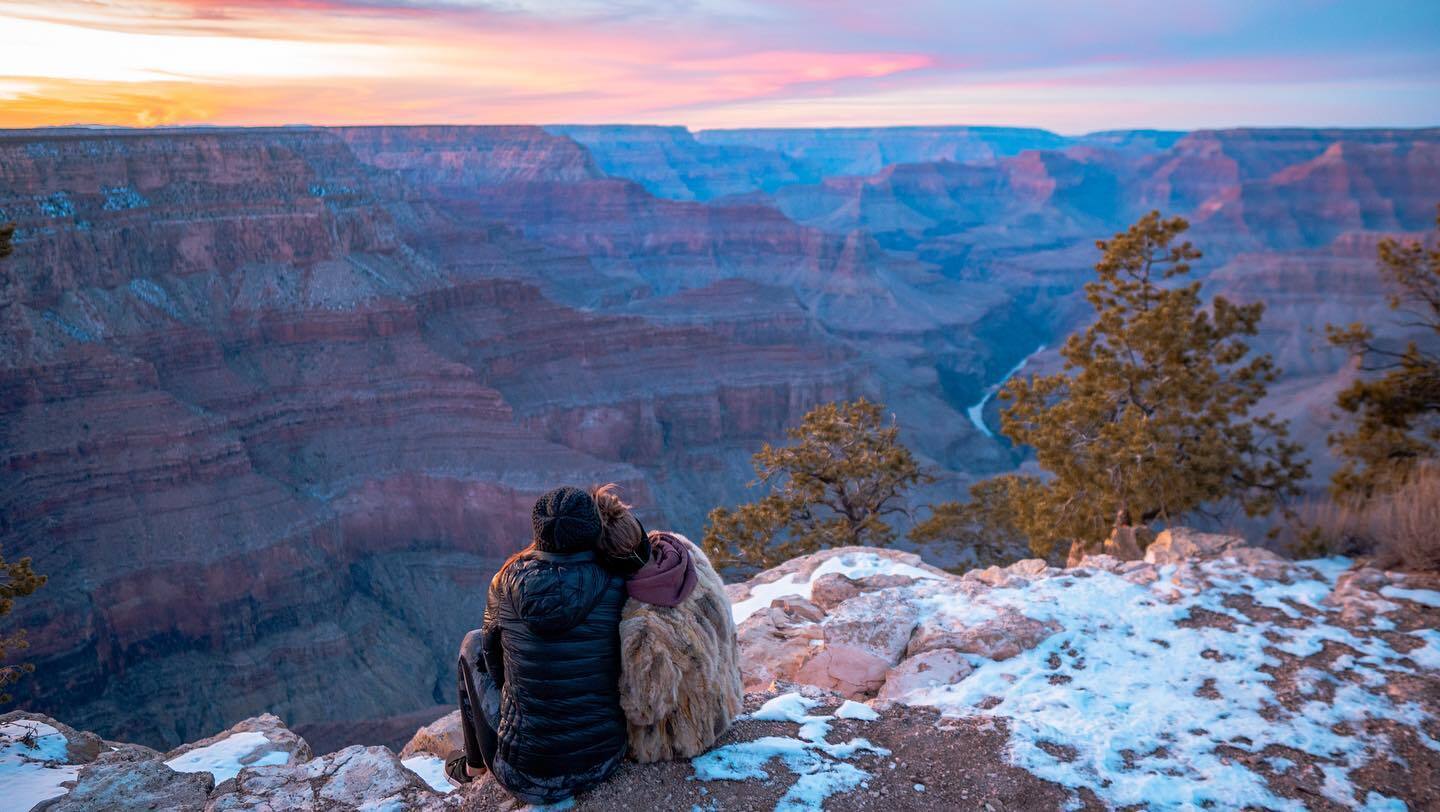 A couple takes in the beautiful snowy sunset at Grand Canyon while sitting and hugging.