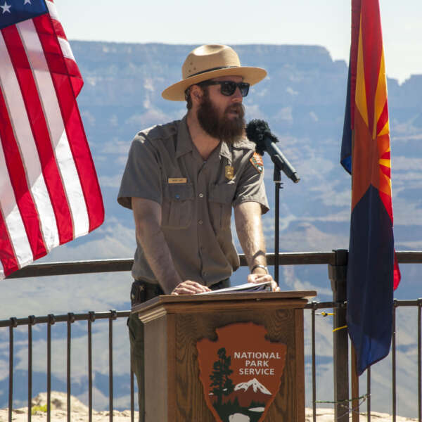 Grand Canyon National Park Ranger, Rader Lane, giving a summary of the park's Dark Sky program. NPS/Bryan Maul (He is wearing a NPS uniform and is standing in between two flags, with a canyon landscape in the background.)